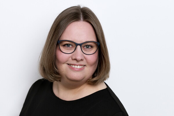 Porträt von Katrin Floß. Sie trägt eine dunkle Brille und ein schwarzes Oberteil. Ihr braunes, kinnlanges Haar ist glatt gestylt. Sie lächelt in die Kamera vor einem hellen Hintergrund.